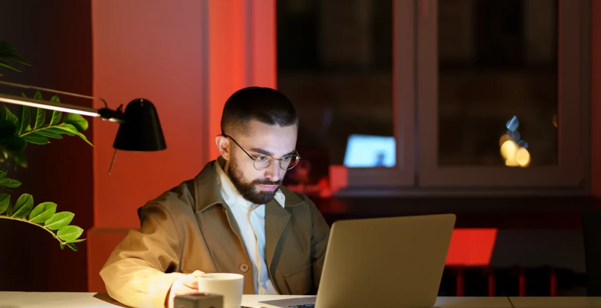 Hombre con gafas concentrado ante un portátil y una taza de café, trabajando de noche