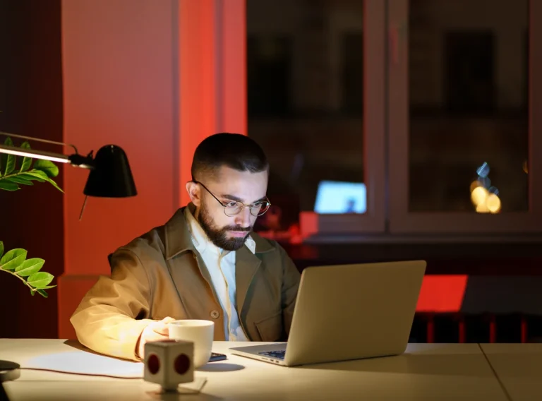 Hombre con gafas concentrado ante un portátil y una taza de café, trabajando de noche Hombre con gafas concentrado ante un portátil y una taza de café, trabajando de noche