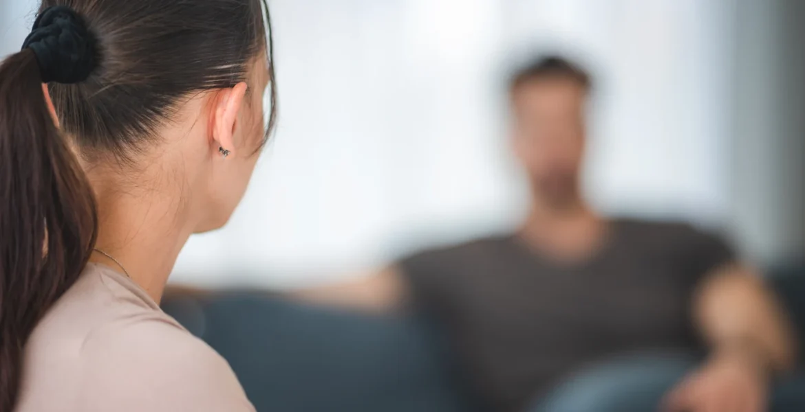 Mujer de espaldas frente a su pareja, sentado al fondo en el sofá